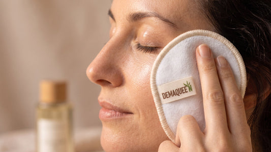 Gros plan sur le profil d'une femme aux yeux fermés se démaquillant délicatement la joue avec un cotondé maquillant réutilisable blanc bordé de beige, étiqueté « DEMAQUEEN ».